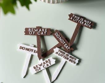 May include: A collection of decorative wooden signs with humorous messages. The signs are in brown and white, with text that reads "Beware of Cat", "Do Not Eat", and "This is Not a Litter Box".