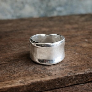 May include: A wide, silver-colored ring with a smooth, rounded exterior. The ring has a thick band and a slightly curved top, resting on a wooden surface. The ring's design is simple and modern, reflecting light.