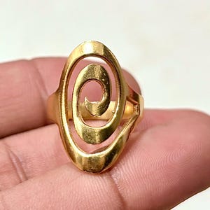 May include: A gold-coloured ring with an oval face featuring a spiral design. The ring is made of a shiny metal and has a simple, elegant style. The ring is shown on a hand.