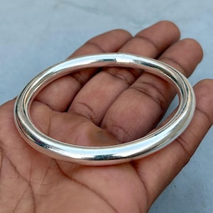 May include: A shiny, silver-colored bangle bracelet. The bracelet is a solid, rounded band. The surface of the bracelet reflects light, creating highlights and shadows. The bangle is a simple, elegant piece of jewelry.