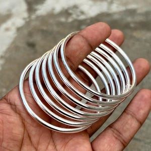 May include: A set of silver bangles, held in a hand. The bangles are thin, circular, and made of metal. Light reflects off the smooth surface, creating highlights. The background is a blurred, neutral tone. The bangles are likely to be worn as jewellery.