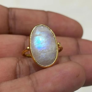 May include: A gold-toned ring featuring an oval, iridescent moonstone. The moonstone displays a play of blue and white colors. The ring has a simple, elegant design, with a slender band and decorative accents on the sides of the stone.
