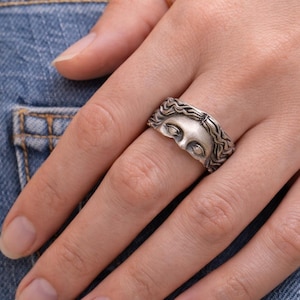 Peut inclure: Bague argentée représentant un visage sculpté avec des yeux fermés et des cheveux ondulés. La bague est portée sur un doigt, avec une simple bague dorée également visible. Le fond est en tissu denim.