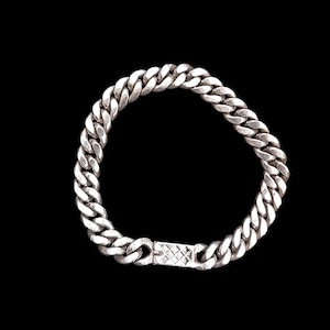 以下が含まれることがあります： 銀色のカーブチェーンブレスレット、長方形の留め金付き。 ブレスレットは太く丸みを帯びたリンクでできており、黒い背景に表示されています。 留め金は、テクスチャ加工されたクロスハッチデザインです。