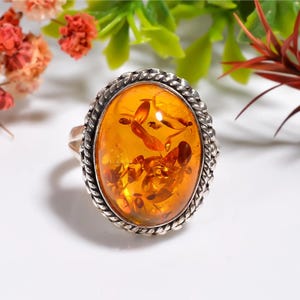 May include: Oval amber ring with a silver-colored, braided border. The amber stone has a warm, orange hue with visible inclusions. The ring is set against a white background with hints of greenery and red flowers.