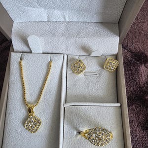 Puede incluir: Un conjunto de joyas doradas presentado en una caja blanca. El conjunto incluye un collar con un colgante cuadrado, pendientes a juego y un anillo. Todas las piezas están incrustadas con pequeñas piedras preciosas transparentes, creando un efecto brillante. La caja está abierta.