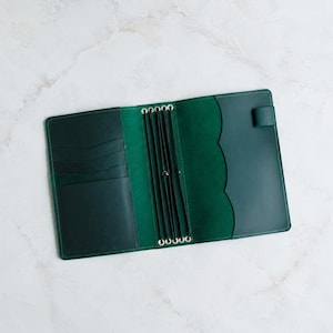 May include: A dark green leather traveler's notebook cover featuring an open interior with multiple card slots on the left side, a central spine with four black elastic bands for securing inserts, and a pen loop on the right side edge.