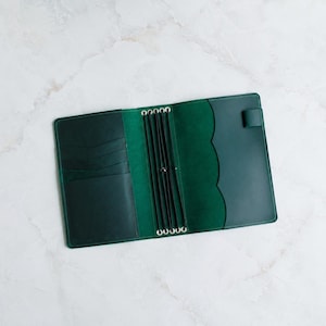 May include: A dark green leather traveler's notebook cover featuring an open interior with multiple card slots on the left side, a central spine with four black elastic bands for securing inserts, and a pen loop on the right side edge.