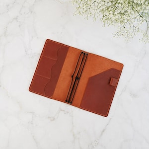 May include: A tan leather notebook cover featuring an open interior with three vertical card slots on the left side, a large pocket on the right, and three black elastic cords running vertically through the center to hold paper inserts.