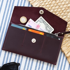 May include: A burgundy leather wallet with multiple compartments, including a zippered section and card slots. The wallet is open, revealing currency and cards. Black sunglasses rest near the wallet on a striped surface.