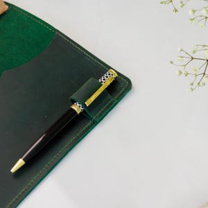Op de afbeelding: Een donkergroene lederen notitieboekhoes met een bijpassende lederen penlus. Een zwarte balpen met een goudkleurige clip en een zwart-wit patroon aan de bovenkant is in de lus gestoken. De hoes heeft zichtbare groene stiksels langs de rand.