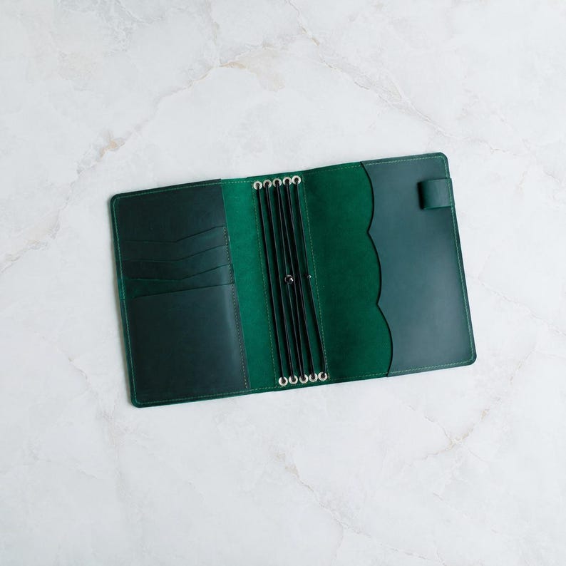 May include: A dark green leather traveler's notebook cover featuring an open interior with multiple card slots on the left side, a central spine with four black elastic bands for securing inserts, and a pen loop on the right side edge.