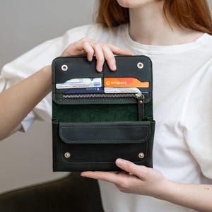 Könnte beinhalten: Dunkelgrüne Ledergeldbörse mit mehreren Fächern, darunter Kartenfächer und eine Reißverschlusstasche. Die Geldbörse ist geöffnet und zeigt das Innere und den Inhalt. Das Design weist sichtbare Nähte und Metall-Druckknöpfe auf. Die Geldbörse wird von einer Person offen gehalten.