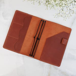 May include: Open brown leather journal with multiple pockets and black elastic closures. The journal is open, revealing a lighter brown interior. The cover has a dark brown pocket and a flap closure. The journal is on a white marble surface.
