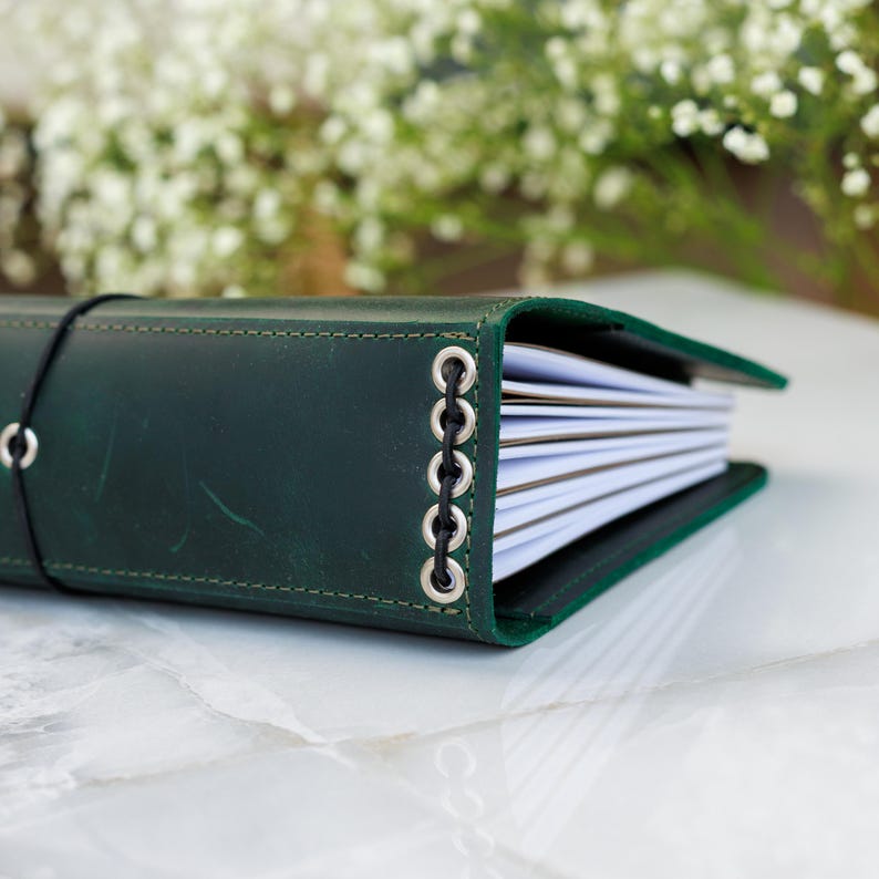 Op de afbeelding: Een donkergroen lederen dagboek met een zwarte elastische sluiting en een zijrug versterkt met vijf zilverkleurige metalen oogjes en een zwart koord, met meerdere witte papieren inlegvellen die aan de zijkant zichtbaar zijn.