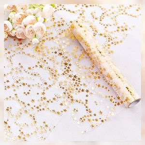 May include: A roll of gold star-patterned wrapping paper. The paper is transparent with a repeating pattern of gold stars. The roll is partially unrolled, revealing the star design. A bouquet of white flowers is in the background.