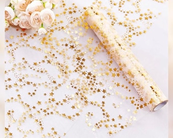 May include: A roll of gold star-patterned wrapping paper. The paper is transparent with a repeating pattern of gold stars. The roll is partially unrolled, revealing the star design. A bouquet of white flowers is in the background.