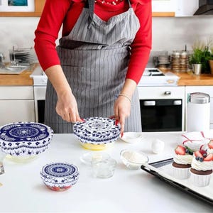 May include: A person is shown in a kitchen setting, placing a blue and white patterned bowl cover on a bowl. Several other bowls with similar covers are visible, along with ingredients and cupcakes. The covers have a floral design.