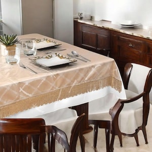 May include: A dining table set for a meal, featuring a beige tablecloth with a diamond pattern and fringe trim. The table is set with square plates, silverware, and glasses. Wooden chairs surround the table, creating an elegant dining setting.