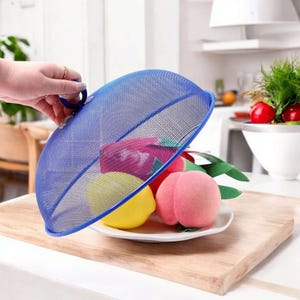 May include: A blue mesh food cover is lifted to reveal a plate of colorful fruit, including a yellow lemon and a pink peach. The cover sits on a wooden cutting board. A hand is lifting the cover. The background shows a kitchen setting.
