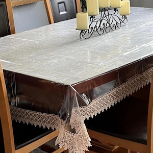 May include: A rectangular dining table covered with a clear plastic tablecloth. The tablecloth has a decorative lace trim. A black metal candelabra with five lit candles sits on the table. The table is surrounded by black chairs.