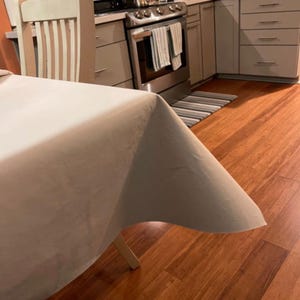 May include: A light beige tablecloth draped over a white wooden table. The table is set in a kitchen with a stainless steel oven and light gray cabinets. The floor is hardwood.
