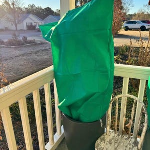 May include: A tall, dark gray planter with a bright green plant cover. The cover is draped over the plant, protecting it from the elements. A distressed white chair sits nearby on a porch with a white railing.