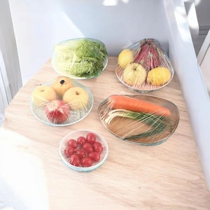 May include: An assortment of fresh produce, including apples, lemons, a carrot, and grapes, wrapped in clear plastic wrap. The items are arranged on small plates and bowls, showcasing a food storage concept.