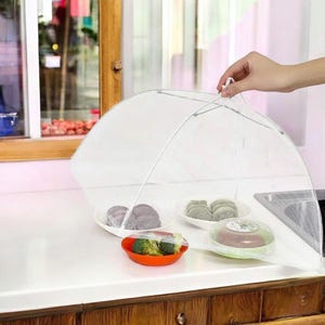 May include: A white, mesh food cover, shaped like an umbrella, protects various dishes on a white countertop. The dishes include a red bowl with broccoli, and plates with food. The cover is held open by a hand.
