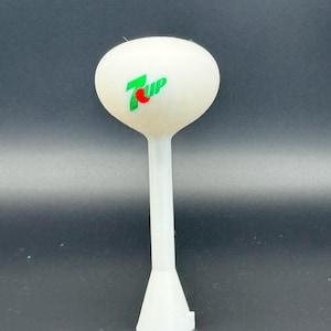 Op de afbeelding: Een wit 3D-geprint model van een watertoren met een lange steel en een uitlopende basis. De bovenkant is voorzien van het 7 Up-logo in groen en rood. Het model staat tegen een donkere achtergrond.
