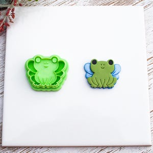 May include: Two frog-shaped items. One is a green plastic frog-shaped cookie cutter. The other is a green frog with blue wings. Both items are on a white surface with a wooden background.