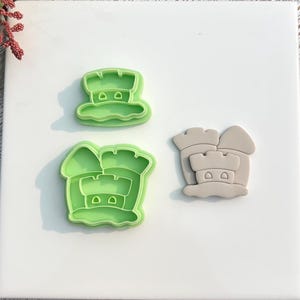May include: Three cookie cutters in the shape of a castle. Two are bright green, and one is a light tan color. The cutters are on a white square tile. The castle design includes turrets and windows.