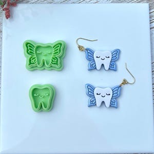 May include: A white square tile displays two green tooth-shaped cookie cutters and two pairs of blue and white tooth fairy earrings with gold-colored hooks. The earrings and cutters have a smiling tooth design with butterfly wings.