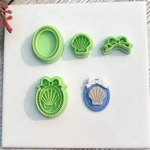 Op de afbeelding: Een verzameling groene koekjessnijders in de vorm van een zeeschelp en een strikvormige snijder. Eén snijder is een strik, de andere zijn ovaal, zeeschelp en zeeschelp met een strik. De snijders liggen op een wit oppervlak.