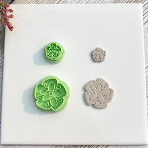 Könnte beinhalten: Vier blütenförmige Ausstecher aus Polymer Clay in verschiedenen Größen. Zwei sind grün und zwei sind beige. Die Ausstecher sind auf einer weißen Oberfläche angeordnet. Die grünen Ausstecher haben einen erhöhten Rand, während die beigen flach sind.
