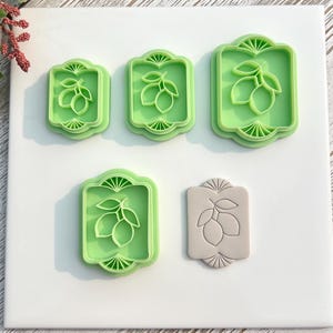 May include: Five green plastic lemon-shaped cookie cutters in various sizes, arranged on a white surface. One beige cookie is also present. The cutters feature a lemon and leaf design within a decorative frame, ideal for baking.