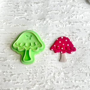 May include: Two mushroom-shaped items. One is a light green cookie cutter with a butterfly detail. The other is a red mushroom with white polka dots and a tan stem. Both are on a white textured surface.