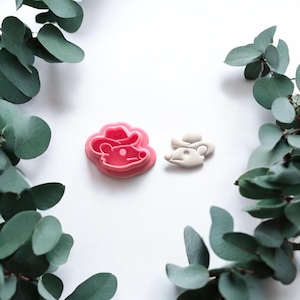 Puede incluir: Un cortador de galletas rosa con forma de ratón y una galleta pequeña con forma de ratón de color claro. El ratón lleva un sombrero de vaquero. El cortador de galletas es de plástico rosa. Hojas verdes rodean los objetos.