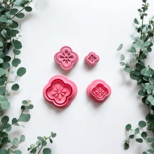 May include: Four pink flower-shaped cookie cutters of varying sizes. The cutters have a four-petal design with a detailed center. The cutters are arranged on a white surface, with green eucalyptus branches in the background.