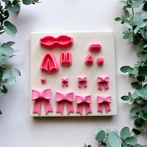 May include: A collection of pink bow-shaped cookie cutters in various sizes, arranged on a cream-colored surface. The cutters are made of plastic and are designed for creating decorative elements for baked goods. The image is well-lit, showcasing the details of each cutter.