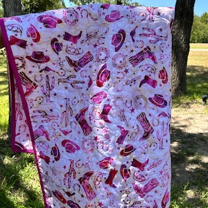 May include: A white quilt with a repeating pattern of cowboy boots, hats, stars, and other western-themed designs in shades of pink and purple. The quilt has a pink border and is held up outdoors.