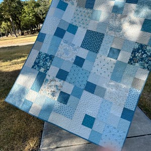 May include: A patchwork quilt featuring a variety of square fabric blocks in shades of blue, light blue, and white. The quilt displays an assortment of patterns including floral prints, geometric textures, and solid colors with intricate stitching.