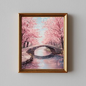 以下が含まれることがあります： ピンク色の桜が咲き誇る木々に囲まれた、川にかかる石橋を描いた額入りの絵画。 金色のフレームに入ったこの作品は、柔らかな筆致で穏やかな風景を描いています。