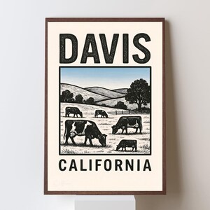 May include: A framed print featuring a pastoral scene with cows grazing in a field under a blue sky. The print includes the text "DAVIS" above and "CALIFORNIA" below the illustration, all in a bold, black font.