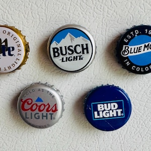 May include: A collection of five beer bottle caps. The caps feature various brand logos, including Miller Lite, Busch Light, Blue Moon, Coors Light, and Bud Light. The caps are in various colors, including gold, silver, blue, and white.