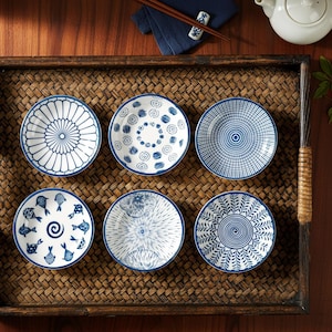 Peut inclure: Six petites assiettes rondes en céramique avec des motifs bleus et blancs sont disposées sur un plateau tressé. Chaque assiette présente un design unique, notamment des motifs floraux, géométriques et de poissons. Les assiettes ont un bord bleu.