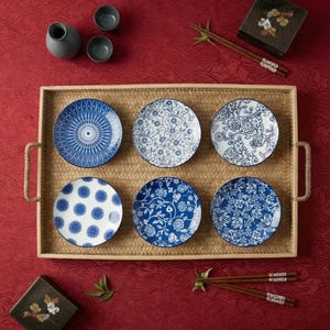 May include: A set of six blue and white patterned ceramic plates arranged on a woven tray. The plates feature various designs, including floral and geometric patterns. Chopsticks and small serving dishes are also present.