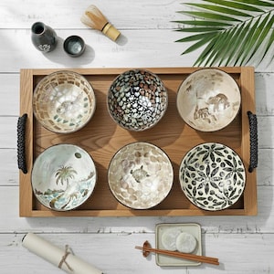 Puede incluir: Una bandeja de madera con seis cuencos decorativos, cada uno con diseños únicos de mosaico o pintados. Los cuencos presentan imágenes de peces, tortugas, elefantes y motivos florales. Artículos adicionales incluyen una pequeña botella de cerámica, una taza de té, un batidor y un plato pequeño con comida.