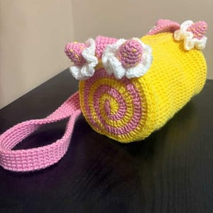 May include: A handmade crochet bag in a roll shape, with a yellow body and pink and yellow spiral detailing on the ends. The bag is decorated with pink and white crochet flowers and has a pink strap.