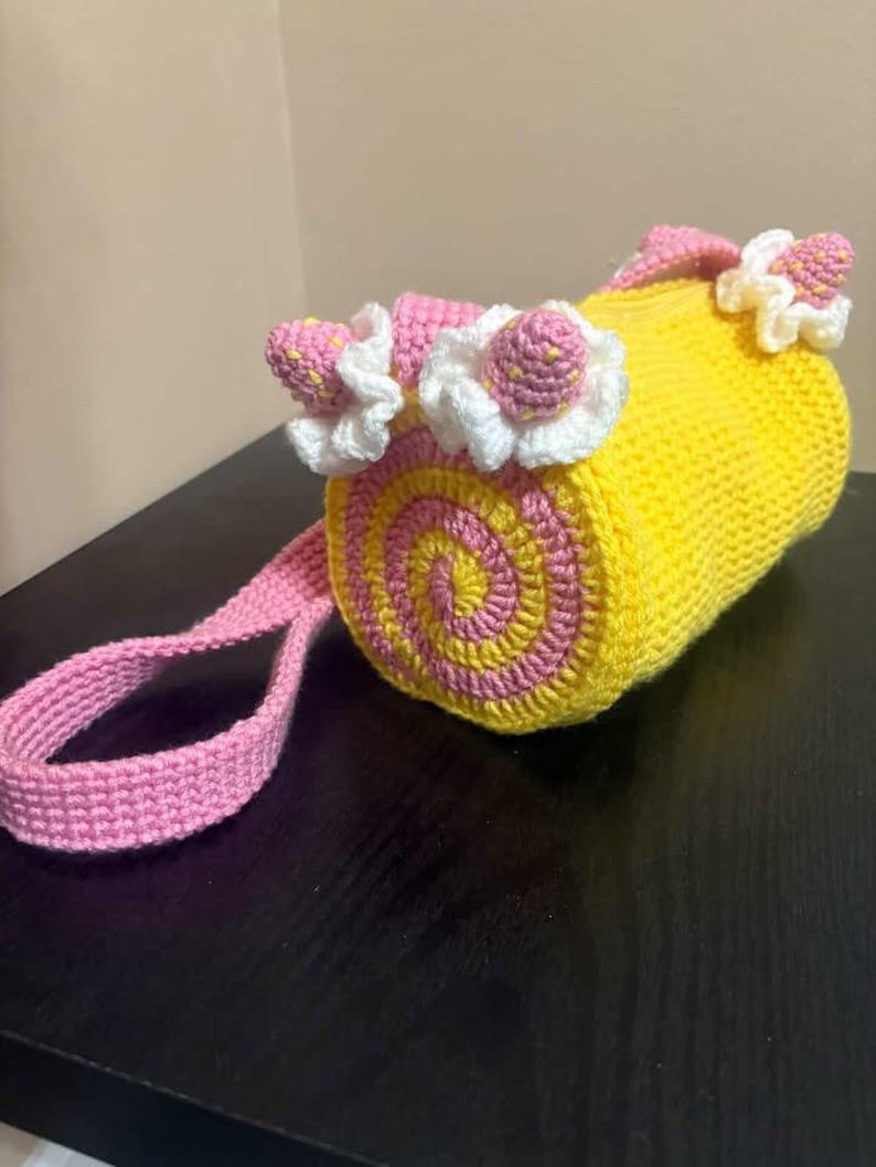 May include: A handmade crochet bag in a roll shape, with a yellow body and pink and yellow spiral detailing on the ends. The bag is decorated with pink and white crochet flowers and has a pink strap.